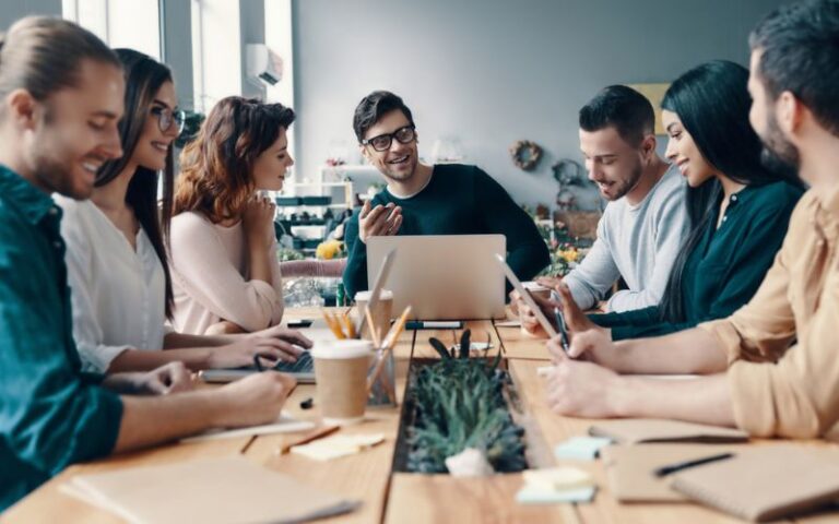 Team of digital marketers collaborating around a table with laptops and smartphones, discussing affiliate marketing strategies and email campaign tools.